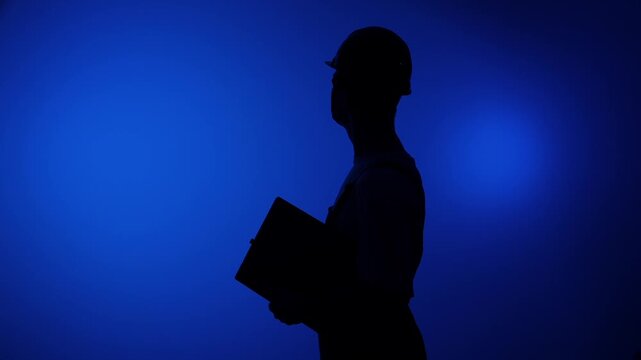 Professional person working in engineering or construction industry, wearing hardhat and holding a clipboard, standing in silhouette against a clear blue background