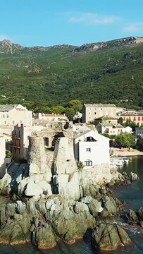 Aerial view of historic stone ruins and traditional buildings along the rocky coastline of Erbalunga, Corsica, with lush green hills in the background. Clear blue sky and tranquil seaside atmosphere.