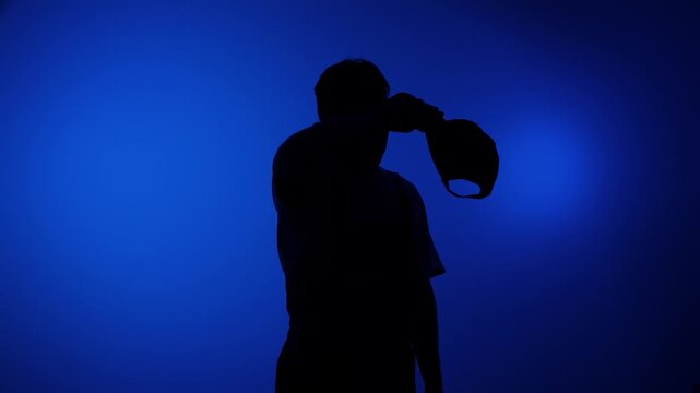 Man in profile creates a striking black silhouette against a vivid deep blue studio backdrop, holding a baseball cap over his head moody, minimal, graphic, and anonymous