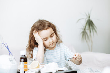 little sick girl sitting on a bed in the bedroom and measures her temperature with a thermometer
