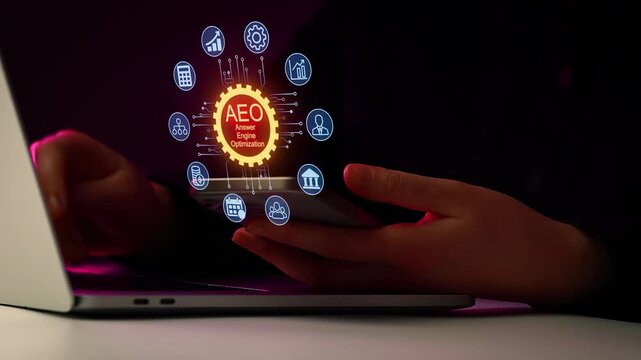 Hands holding a smartphone while using a laptop to explore AEO for better search engine visibility at night.