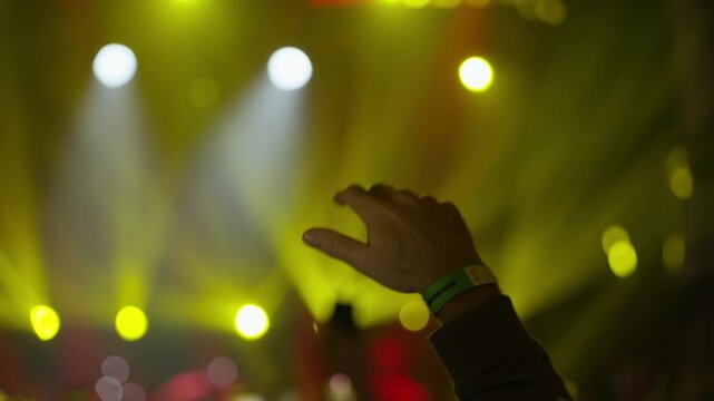 An energetic concertgoer's hand sways with green wristbands against a backdrop of vibrant, changing stage lights, creating an immersive live music experience. Dynamic lighting and crowd atmosphere.