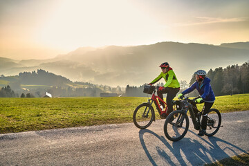 Fototapeta premium Active senior couple riding electric bikes through scenic Bregenzerwald in Austria during golden evening light. Enjoying nature, mountains, and healthy outdoor lifestyle in spring countryside. 