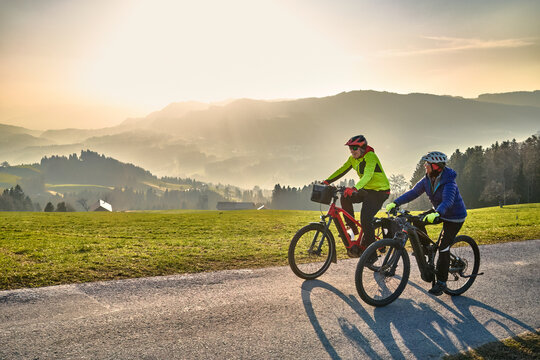 Active senior couple riding electric bikes through scenic Bregenzerwald in Austria during golden evening light. Enjoying nature, mountains, and healthy outdoor lifestyle in spring countryside.
