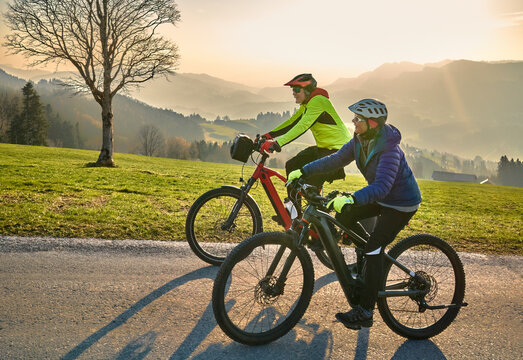 Active senior couple riding electric bikes through scenic Bregenzerwald in Austria during golden evening light. Enjoying nature, mountains, and healthy outdoor lifestyle in spring countryside.
