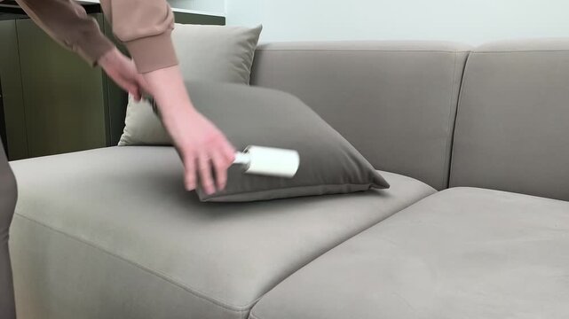 Hand cleans cushion with lint roller removing fur and dust. Person rolls adhesive tool over velour upholstery at home. Cleaning process and hygiene maintenance concept in apartment living room..