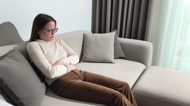 Woman sits on sofa holding stomach from sudden pain. Female touches abdomen feeling discomfort and internal tension. Condition suggests digestive issue and need for attention to health..