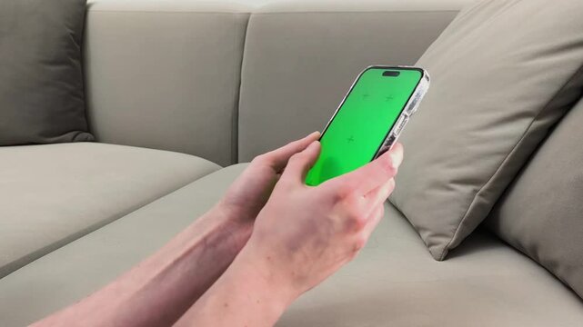 Hands hold smartphone and swipe left on green screen while sitting near sofa at home. Fingers perform gesture across chroma display device with touch interaction. Concept digital interface