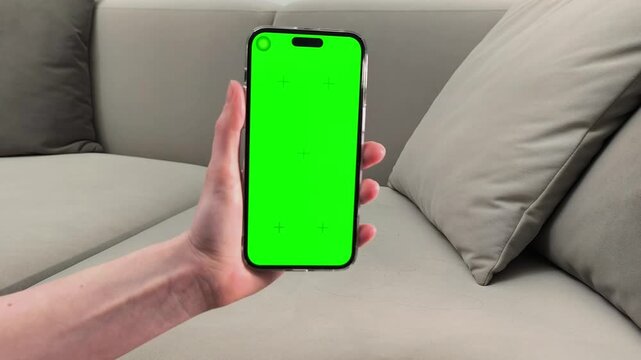 Hand holds smartphone with green screen and taps lower part before scrolling interface. Finger presses and swipes on chroma display device at home. Concept app navigation, digital