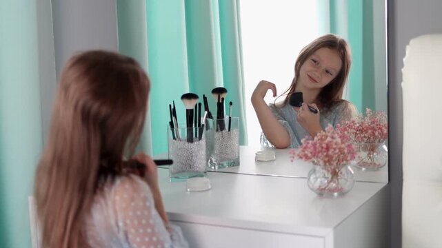 A smiling little girl pretending to do makeup at a vanity mirror, holding a cosmetic brush. Child play, imitation of beauty routines, innocence, self-expression, and creativity at home. 4k footage