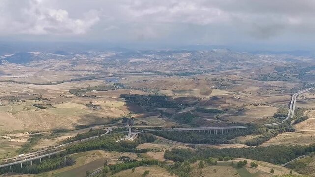 Panoramic view of rolling hills and valleys in the Province of Enna, Sicily, showcasing a winding road and lush greenery under a cloudy sky