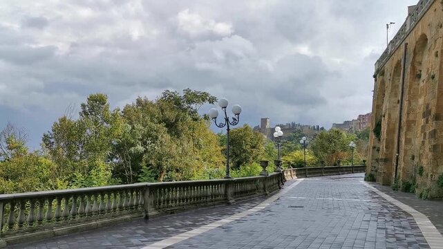 Scenic view of a winding street lined with trees and lampposts, leading towards Lombardy Castle in Enna, Sicily, under a cloudy sky with a hint of sunlight