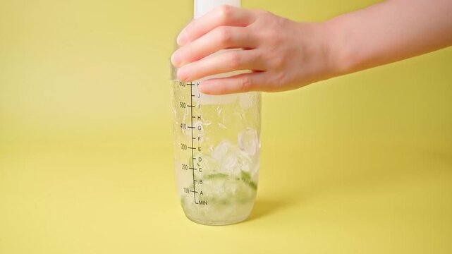 Hand shaking clear bottle making refreshing iced smashed lime tea with fresh green lime slices and ice cubes on bright yellow background macro shot