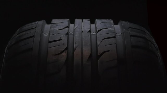 Slow Rotation of the Tire in the Dark. The new car tire is highlighted by a spotlight in the dark. The wheel rotates slowly toward the viewer against a black background