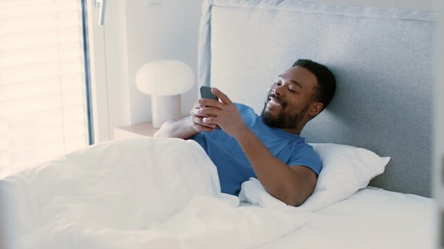 A man with dark skin is lying in bed under white covers. He is smiling and looking at his smartphone. The setting is bright and cozy, indicating a relaxed morning atmosphere.