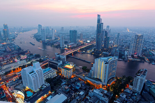 Aerial panorama of Bangkok in evening twilight with busy traffic trails on Taksin Bridge, light trails of boats on Chao Phraya River & skyscrapers by riverside ~ Night in Bangkok in bird's eye view