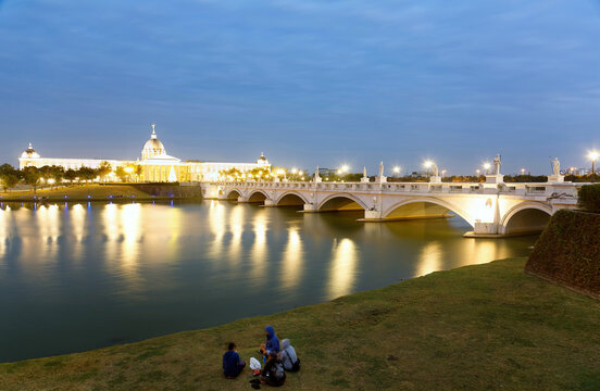 Tourists sitting on the riverside meadow & enjoying the scenery of Chimei Museum at blue hour, with the pseudo-classical architecture & bridge illuminated & reflected in the water, in Tainan, Taiwan