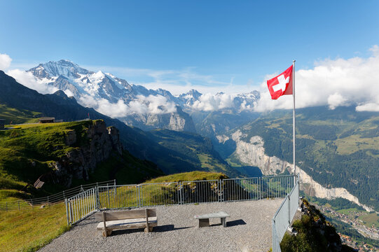 Swiss flag waving on a viewpoint at the edge of a cliff with snow capped Jungfrau in background & Lauterbrunnen village in the valley on a sunny summer day in Mannlichen, Bernese Oberland, Switzerland
