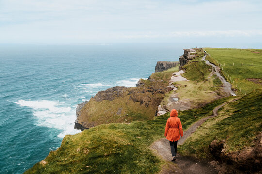 A person in a bright orange jacket walks along a narrow coastal path winding high above the churning blue waters of the Atlantic at the Cliffs of Moher.