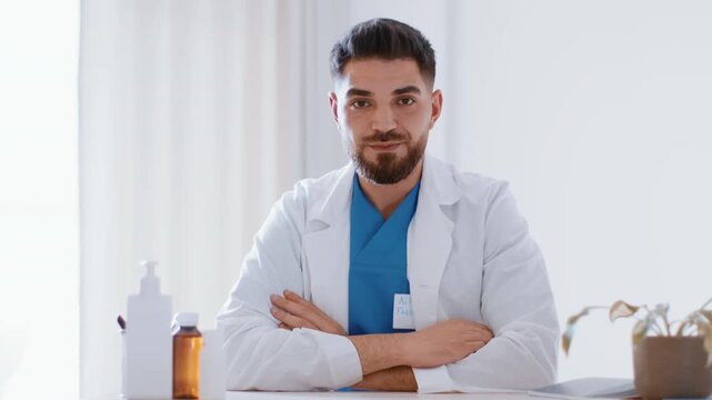 A man with a beard wearing a white coat and blue scrubs sits at a desk in a medical office. He has his arms crossed and looks directly at the camera. Several healthcare items are on the desk.