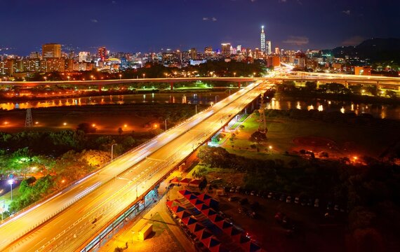 Aerial view of Fuhe Bridge spanning Xindian River & an expressway passing over Keelung Road, with 101 Tower standing among crowded buildings in Xinyi District & city lights dazzling at night in Taipei