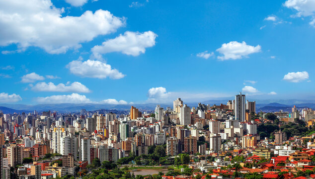 Vista panor&acirc;mica da vibrante regi&atilde;o Centro-Sul de Belo Horizonte, Brasil, exibindo um horizonte denso de edif&iacute;cios coloridos contra um pano de fundo de montanhas onduladas e um c&eacute;u azul brilhante.
