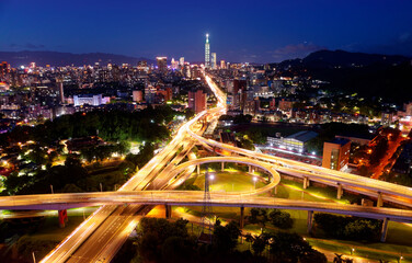 Obraz premium Aerial view over the circular interchange of Shuiyuan Expressway, Keelung Road & Fuhe Bridge in Taipei, with 101 Tower standing amid crowded buildings in Xinyi District & city lights dazzling at dusk