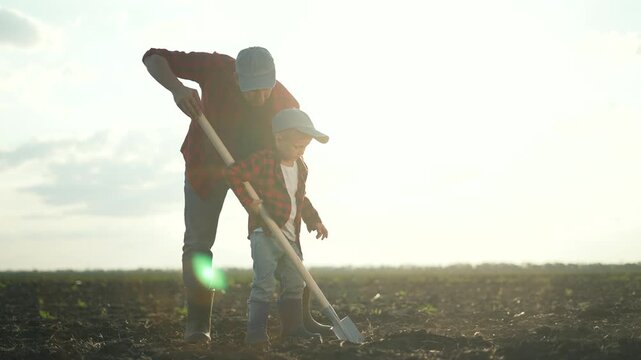 Father teaching son farming. Agriculture farmer showing son how to use shovel field. Father and son bonding while farming. Agriculture education rural setting. Farmer teaches son in agriculture field.