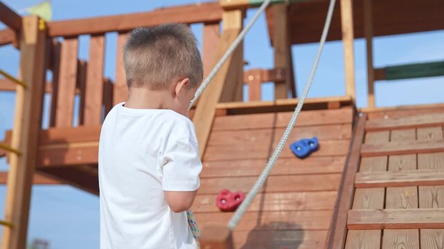 Young Child climbing rope at playground. Child plays on wooden play structure in sunny weather. Happy childhood outdoors. Child pulling rope at playground. Active child play, enjoying playground fun.