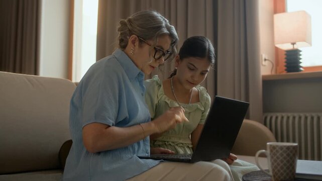 Medium long shot of young granddaughter teaching grandmother typing on keyboard of wireless laptop while sitting on couch in living room
