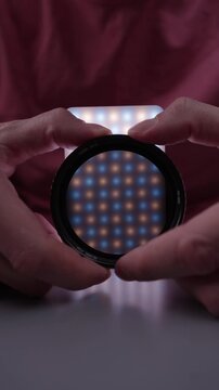 Hands holding rotating circular polarizing filter, adjusting effect on bicolor led light panel for photography and video, demonstrating light reflection reduction, close-up, vertical shot, slowmo.