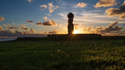 Moai statue silhouette stands peacefully on Easter Island under breathtaking sunset sky representing mysterious ancient Rapa Nui culture with bright golden sun ray over green grass field © Jirawatfoto