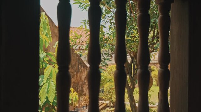 Looking out from a dim interior through ornate dark timber window balustrades to a vibrant sunny backyard garden featuring green foliage and terracotta roof tiles in daylight.
