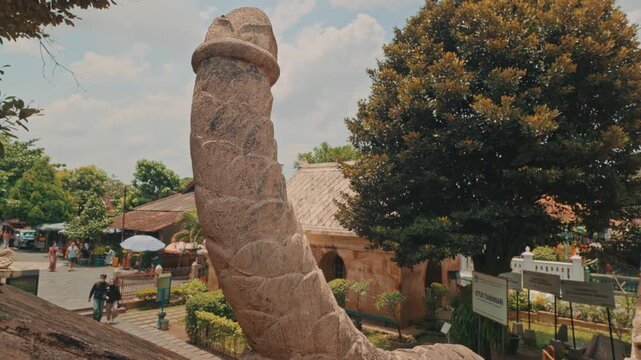 A stone statue shaped like a phallic symbol stands prominently against a bright blue sky at an outdoor historic landmark or park, capturing an exotic architectural detail.