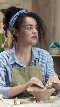 Curly woman wearing apron shaping clay bowl and looking aside. Female pressing material with fingers and forming edges. Thinking about design and continuing work. Working in workshop.