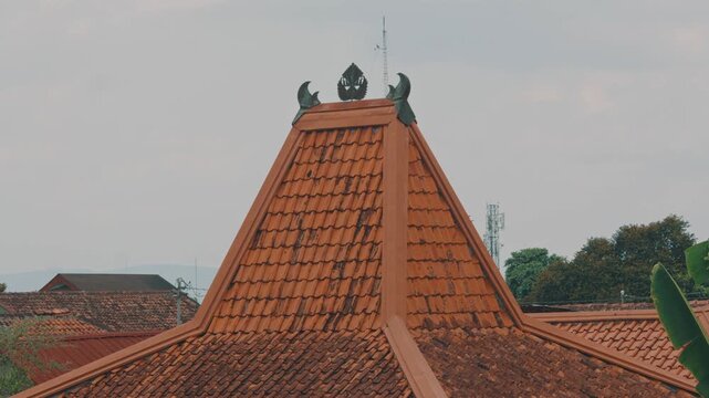 A low angle view of an ornate, terracotta-tiled traditional Indonesian Joglo house roof against a soft overcast sky, featuring a decorative wooden peak ornament.