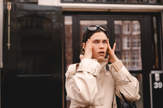 Annoyed young woman rolling eyes and holding hands to head on city street, frustrated female traveler in beige trench coat showing irritation, negative emotion and stress concept