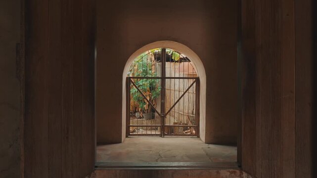 An elegant symmetrical perspective looking through a rustic arched stone entryway into a sunlit courtyard garden with lush greenery and a vintage metal gate.