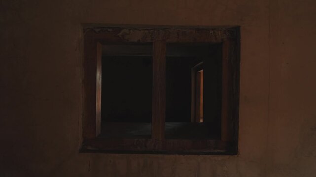 A low-light, atmospheric shot looking through a rustic window frame into a dark, abandoned room, evoking a sense of mystery, stillness, and historical decay in a moody space.