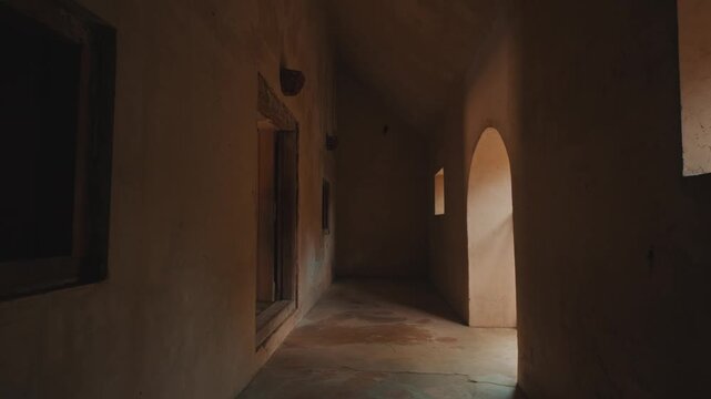 A moody perspective shot moving through a narrow, sunlit corridor inside a weathered historical stone structure with earthy textures and dramatic architectural lighting shadows.