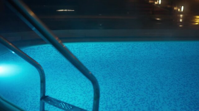 A tranquil swimming pool at night, illuminated by a glowing blue underwater light, creating a peaceful and serene atmosphere. Steel ladders offer entry into the clear, mosaic-tiled water,