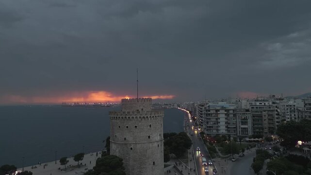 Cinematic aerial view of the White Tower in Thessaloniki during a dramatic sunset. Fiery sun piercing through dark heavy clouds over the Thermaic Gulf, Greece.