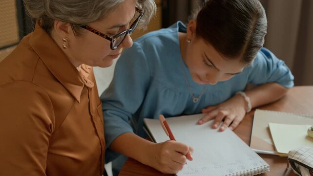 Handheld shot of Caucasian grandma assisting young girl writing in notebook while doing homework together
