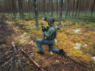 Naklejka premium Military drone pilot in camouflage gear kneeling in a pine forest holding a remote controller with night vision goggles mounted