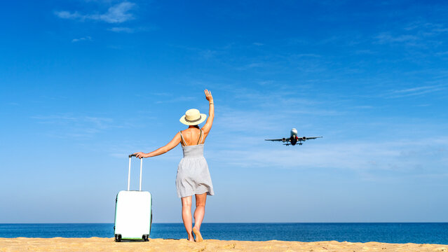Woman waves goodbye from beachside, suitcase beside her