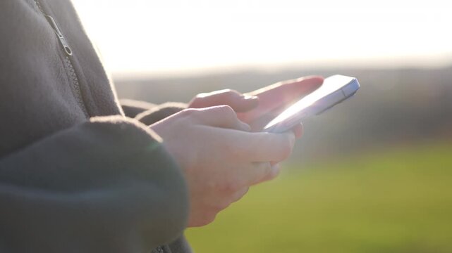 Close up of hands holding smartphone outdoors, sunlight creates soft lens flares on screen, blurred green background, calm and natural everyday moment.