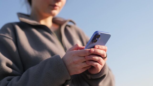 Low angle close up of hands holding blue smartphone against blue sky, grey fleece visible, soft color harmony, woman blurred in background, focus on phone, calm everyday moment.