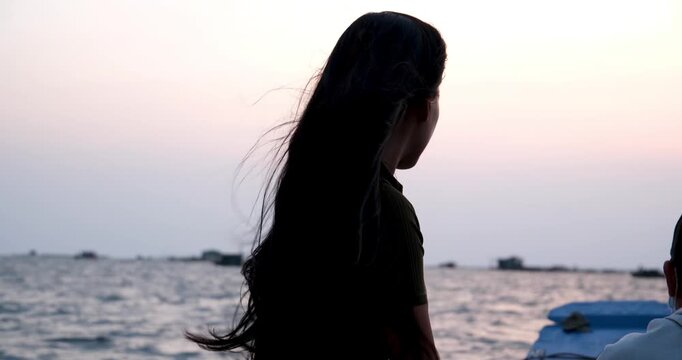 Woman with black braids on boat at sunrise, facing away, wind blowing hair, violet-blue ocean and sky, two other shadowed passengers, mysterious, serene early morning atmosphere.