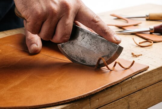 Leatherworker hand using traditional round knife to cut curved shape in brown leather sheet