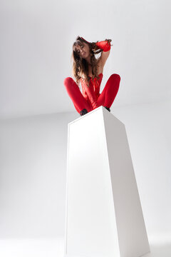 Sexy young woman in red corset and stockings posing on white cube